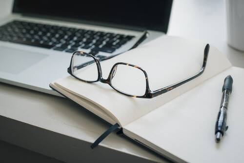 A pair of glasses and a pen on an open notebook next to a laptop